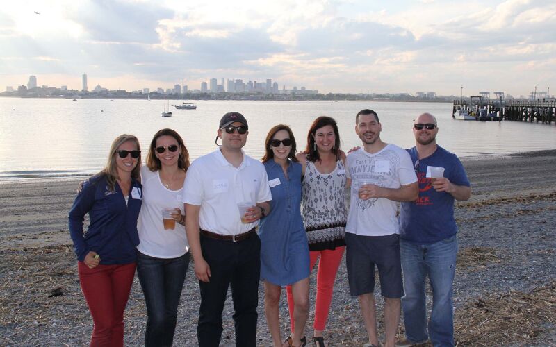 The image shows a group of seven people posing for a photo on a beach. They are all smiling and appear to be enjoying themselves. In the background, there is a body of water and a city skyline. The sky is overcast, suggesting it might be either early morning or late afternoon.
